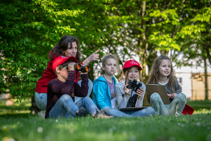Kinder sitzen auf der Wiese und beobachten die Natur mit Fernglas und Notizblock.